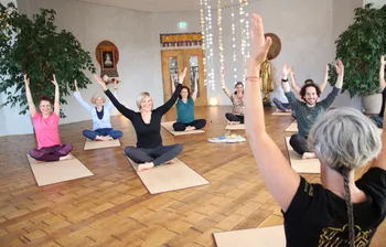 Eine Yoga-Klasse lächelt bei den Übungen die Yogalehrerin an, welche mit dem Rücken zum Betrachter sitzt
